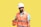 A male construction worker wearing a white hard hat and orange safety vest is holding an orange case. He is looking at the case, standing against a plain yellow background. The worker is also wearing a blue shirt under the vest.