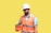 A male construction worker wearing a white hard hat and orange safety vest is holding an orange case. He is looking at the case, standing against a plain yellow background. The worker is also wearing a blue shirt under the vest.
