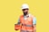 A male construction worker wearing a white hard hat and orange safety vest is holding an orange case. He is looking at the case, standing against a plain yellow background. The worker is also wearing a blue shirt under the vest.