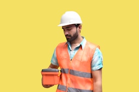 A male construction worker wearing a white hard hat and orange safety vest is holding an orange case. He is looking at the case, standing against a plain yellow background. The worker is also wearing a blue shirt under the vest.