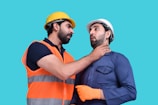 Two men wearing construction helmets are interacting in a manner that appears intense and confrontational. One of them, dressed in an orange safety vest, has his hand around the neck of the other man, who is wearing a blue work uniform and showing a surprised expression.