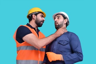 Two men wearing construction helmets are interacting in a manner that appears intense and confrontational. One of them, dressed in an orange safety vest, has his hand around the neck of the other man, who is wearing a blue work uniform and showing a surprised expression.