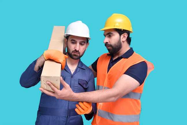 Two men wearing safety helmets and work gloves are observed in a work environment. One man in a white helmet is holding a cardboard box, while the other man in a yellow helmet and an orange safety vest appears to be giving instructions or guidance.