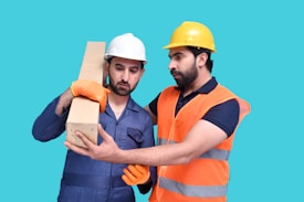 Two men wearing safety helmets and work gloves are observed in a work environment. One man in a white helmet is holding a cardboard box, while the other man in a yellow helmet and an orange safety vest appears to be giving instructions or guidance.