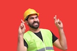 A person wearing a yellow construction helmet and a neon green safety vest is standing against a bright red background with fingers crossed on both hands, expressing hope or good luck.