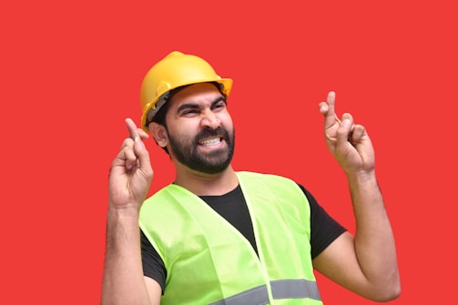 A person wearing a yellow construction helmet and a neon green safety vest is standing against a bright red background with fingers crossed on both hands, expressing hope or good luck.