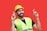 A person wearing a yellow construction helmet and a neon green safety vest is standing against a bright red background with fingers crossed on both hands, expressing hope or good luck.