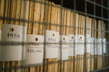 A collection of narrow wooden files organized vertically, each labeled with handwritten tags, indicating the year 1955 and a numerical range, visible through a metal grid.