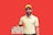 A person wearing a yellow polo shirt and matching cap holds two pizza boxes with a cheerful expression. The background is a solid red color.