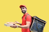 A person is wearing a red polo shirt and cap, carrying a large insulated delivery bag on their back. They hold a pizza box and a paper bag, smiling against a bright yellow background.