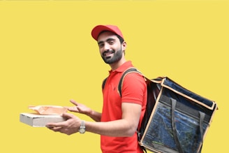 A person is wearing a red polo shirt and cap, carrying a large insulated delivery bag on their back. They hold a pizza box and a paper bag, smiling against a bright yellow background.