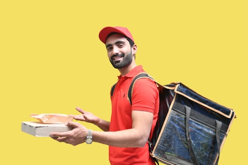 A person is wearing a red polo shirt and cap, carrying a large insulated delivery bag on their back. They hold a pizza box and a paper bag, smiling against a bright yellow background.