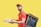 A person is wearing a red polo shirt and cap, carrying a large insulated delivery bag on their back. They hold a pizza box and a paper bag, smiling against a bright yellow background.
