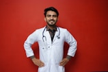 A smiling person in a white lab coat stands confidently against a solid red background, with hands on their hips. A stethoscope hangs around the neck, suggesting a medical professional.