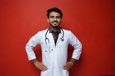 A smiling person in a white lab coat stands confidently against a solid red background, with hands on their hips. A stethoscope hangs around the neck, suggesting a medical professional.