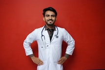 A smiling person in a white lab coat stands confidently against a solid red background, with hands on their hips. A stethoscope hangs around the neck, suggesting a medical professional.