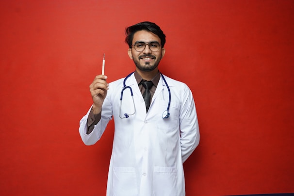 A person wearing a white lab coat and a stethoscope is holding a syringe. The background is a solid red color. The individual is smiling and standing with one hand behind their back.