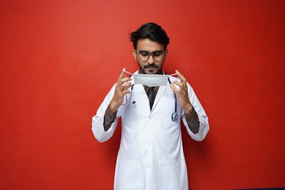 A person wearing glasses dressed in a white lab coat holding a surgical mask against a bright red background. A stethoscope is draped around the neck, suggesting a medical professional.