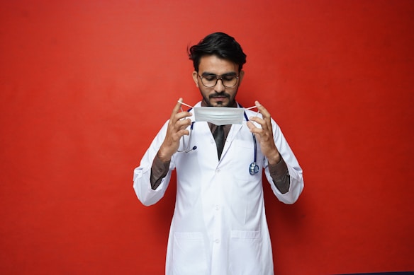 A person wearing glasses dressed in a white lab coat holding a surgical mask against a bright red background. A stethoscope is draped around the neck, suggesting a medical professional.