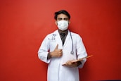 A person wearing a white medical coat and a face mask holds a clipboard and gives a thumbs-up gesture. They have a stethoscope around their neck, and the background is a solid red color.
