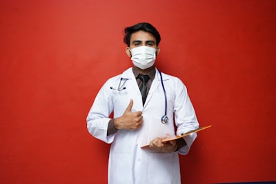 A person wearing a white medical coat and a face mask holds a clipboard and gives a thumbs-up gesture. They have a stethoscope around their neck, and the background is a solid red color.