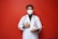 A person wearing a white medical coat and a face mask holds a clipboard and gives a thumbs-up gesture. They have a stethoscope around their neck, and the background is a solid red color.