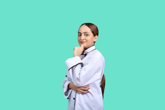 A person in a chef's uniform stands confidently with a hand on their chin. The background is a solid teal color, creating a striking contrast with the white uniform.