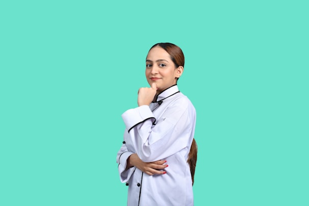 A person in a chef's uniform stands confidently with a hand on their chin. The background is a solid teal color, creating a striking contrast with the white uniform.