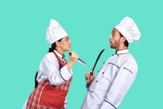Two chefs are in a humorous, dramatic pose where one chef is holding a knife aggressively towards the other, who looks shocked or frightened. Both are wearing traditional chef uniforms, including hats. The background is a solid turquoise color.