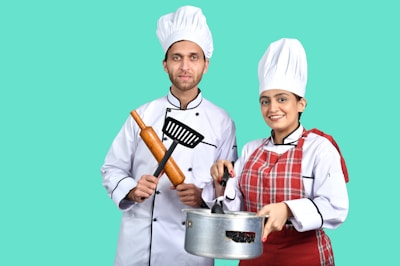 Two chefs in white uniforms and hats stand side by side. The male chef holds a rolling pin and a spatula, while the female chef is smiling and holding a pot with a ladle. She wears a red checkered apron over her uniform. The background is a solid teal color.