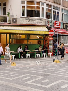 A street scene featuring a small, vibrant café with green exterior walls and white signage, where several people are sitting at tables with white stools, enjoying conversations. The café is located on the ground floor of a building that also houses a law office, indicated by the sign above. Pedestrians walk by, including a woman in a floral dress and a man in a pink shirt. A Coca-Cola vending machine and red signage are visible in the background, along with some greenery.