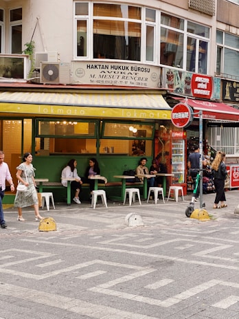 A street scene featuring a small, vibrant café with green exterior walls and white signage, where several people are sitting at tables with white stools, enjoying conversations. The café is located on the ground floor of a building that also houses a law office, indicated by the sign above. Pedestrians walk by, including a woman in a floral dress and a man in a pink shirt. A Coca-Cola vending machine and red signage are visible in the background, along with some greenery.