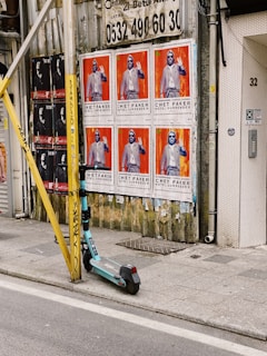 A series of large posters feature a figure in a suit and sunglasses against a vibrant red background. They are plastered on a weathered wall beside a yellow metal pole with graffiti. An electric scooter is parked on the sidewalk with urban surroundings, including wire mesh on a door with the number 32.