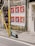 A series of large posters feature a figure in a suit and sunglasses against a vibrant red background. They are plastered on a weathered wall beside a yellow metal pole with graffiti. An electric scooter is parked on the sidewalk with urban surroundings, including wire mesh on a door with the number 32.