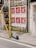A series of large posters feature a figure in a suit and sunglasses against a vibrant red background. They are plastered on a weathered wall beside a yellow metal pole with graffiti. An electric scooter is parked on the sidewalk with urban surroundings, including wire mesh on a door with the number 32.
