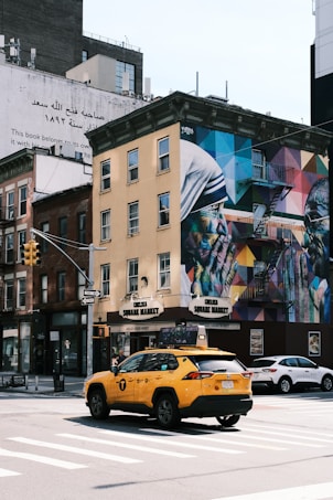 An urban street corner features a yellow taxi in the foreground and a building with vibrant murals painted on its side. The building includes a blend of geometric patterns and colorful portraits. A street sign, traffic lights, and other vehicles are also visible, creating a lively city atmosphere.
