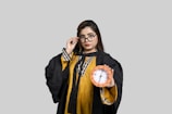 A person wearing a graduation gown and glasses holds a clock in one hand, set against a plain gray background.