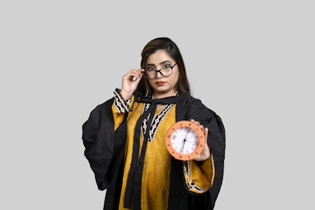 A person wearing a graduation gown and glasses holds a clock in one hand, set against a plain gray background.