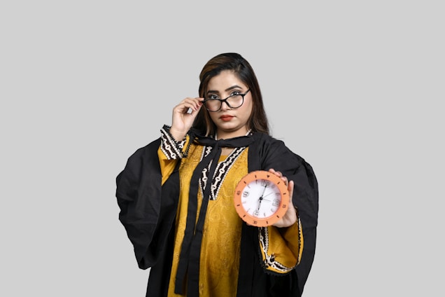 A person wearing a graduation gown and glasses holds a clock in one hand, set against a plain gray background.