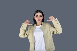 A person with long black hair is wearing a beige jacket and white shirt. She is smiling and pointing both thumbs towards herself while standing against a plain grey background.