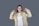 A person with long black hair is wearing a beige jacket and white shirt. She is smiling and pointing both thumbs towards herself while standing against a plain grey background.