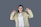 A person with long black hair is wearing a beige jacket and white shirt. She is smiling and pointing both thumbs towards herself while standing against a plain grey background.