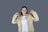 A person with long black hair is wearing a beige jacket and white shirt. She is smiling and pointing both thumbs towards herself while standing against a plain grey background.