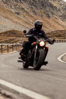 A person wearing a helmet is riding a motorcycle on a winding mountain road, with rocky terrain and a metal railing along the side. The background features a rugged mountain landscape.