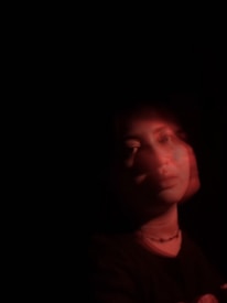 A portrait of a person with a blurred double exposure effect, bathed in red light against a dark background. The face and shoulders are partially visible, creating an ethereal and mysterious atmosphere.