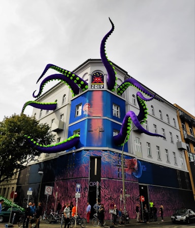 A building features a striking art installation with massive, inflatable purple tentacles with green accents emerging from its windows. The facade is painted with vibrant marine-themed graffiti, showcasing jellyfish and underwater scenes. People are gathered on the sidewalk, some with bicycles.