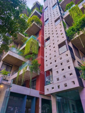 A tall modern building featuring a mix of concrete and red brick with several balconies adorned with green plants. The architecture incorporates geometric patterns and glass elements, creating an eco-friendly and urban aesthetic. Lush greenery cascades from the balconies, adding a natural contrast to the sleek design.