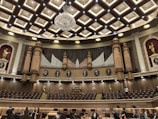 An ornate concert hall features a large pipe organ with decorative woodwork and portraits of composers above. The ceiling displays an intricate pattern with a grand chandelier at its center. Statues of figures in classical poses are placed on the sides. The lower level shows musicians and musical stands, indicating preparations for a performance.