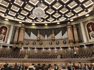 An ornate concert hall features a large pipe organ with decorative woodwork and portraits of composers above. The ceiling displays an intricate pattern with a grand chandelier at its center. Statues of figures in classical poses are placed on the sides. The lower level shows musicians and musical stands, indicating preparations for a performance.