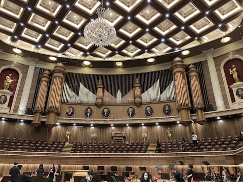 An ornate concert hall features a large pipe organ with decorative woodwork and portraits of composers above. The ceiling displays an intricate pattern with a grand chandelier at its center. Statues of figures in classical poses are placed on the sides. The lower level shows musicians and musical stands, indicating preparations for a performance.
